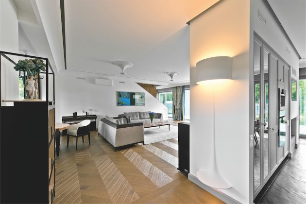 Svetainę nuo virtuvės zonos skiriančią sieną pagyvina priglaistomas minimalistinis šviestuvas „Flos Soft Spun Large“ (britų dizaineris Sebastianas Wrongas). Primenantis toršerą, tarsi pusiau išlindęs iš sienos, neužgožiantis tako, nekliūvantis, jis maloniai nustebina svečius.